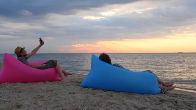 日出时，夫妇坐在海滩上的充气沙发上。年轻人在海滩背景下用日落自拍。男人和女人在假期有放松的时间。心理