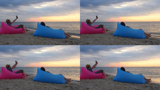 日出时，夫妇坐在海滩上的充气沙发上。年轻人在海滩背景下用日落自拍。男人和女人在假期有放松的时间。心理