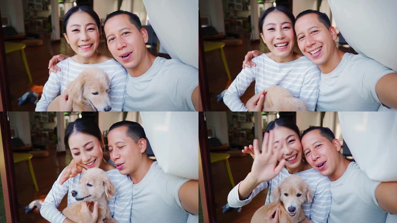 年轻的亚洲成年夫妇抱着一只小狗从手机上自拍，背景是家庭内部。30多岁的成熟男人和女人带着狗宠物拍摄全