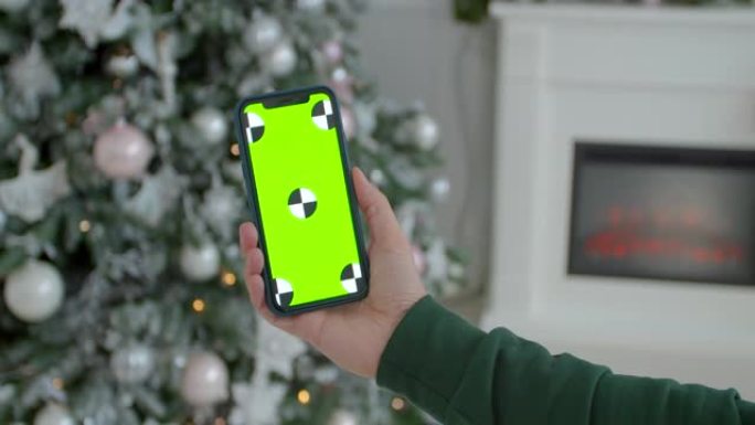 男子在新年装饰的房间里拿着模拟智能手机绿屏铬钥匙。在发光的圣诞家居室内，男性手握黑色现代智能手机的特