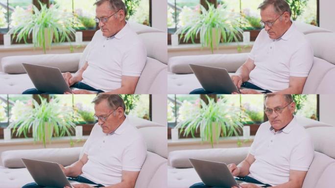 一位退休的老人在家中沙发上的笔记本电脑上打字。年迈的祖父戴着眼镜，在客厅用笔记本电脑工作