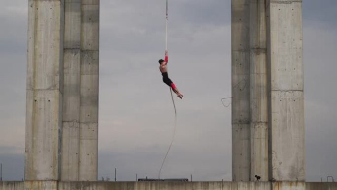强壮肌肉的马戏团艺术家在空中绳索上表演天空背景上的两个混凝土柱之间的户外，勇气、控制和耐力的概念