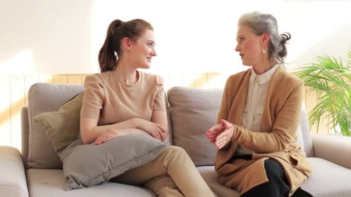 微笑的中年妇女与快乐的成年女儿在沙发上放松，谈论生活时刻。