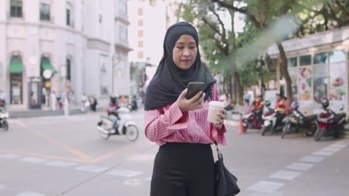 亚洲年轻的穆斯林妇女打电话，拿着咖啡杯站在街边的下午咖啡