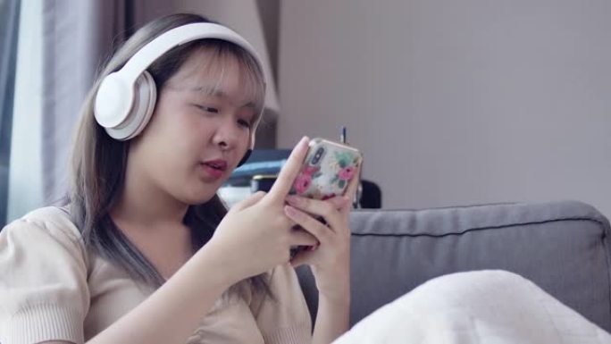 一个亚洲美丽的少女在公寓房间的客厅里用耳机和智能手机听音乐。
