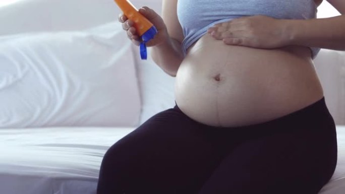年轻的亚洲孕妇母亲坐在床上时拿着乳液瓶和腹部擦乳液