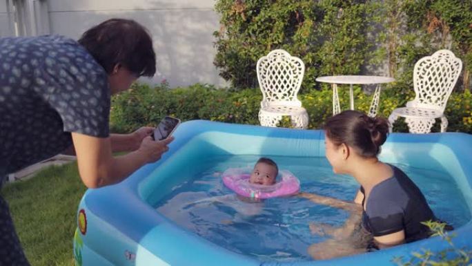 一位祖母正在为在游泳池家庭后院草坪上玩耍的母亲和婴儿拍照