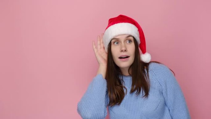 穿着圣诞红色帽子的可爱年轻女子试着听到你偷听，专心听着，穿着舒适的蓝色毛衣，孤立在粉红色背景工作室。