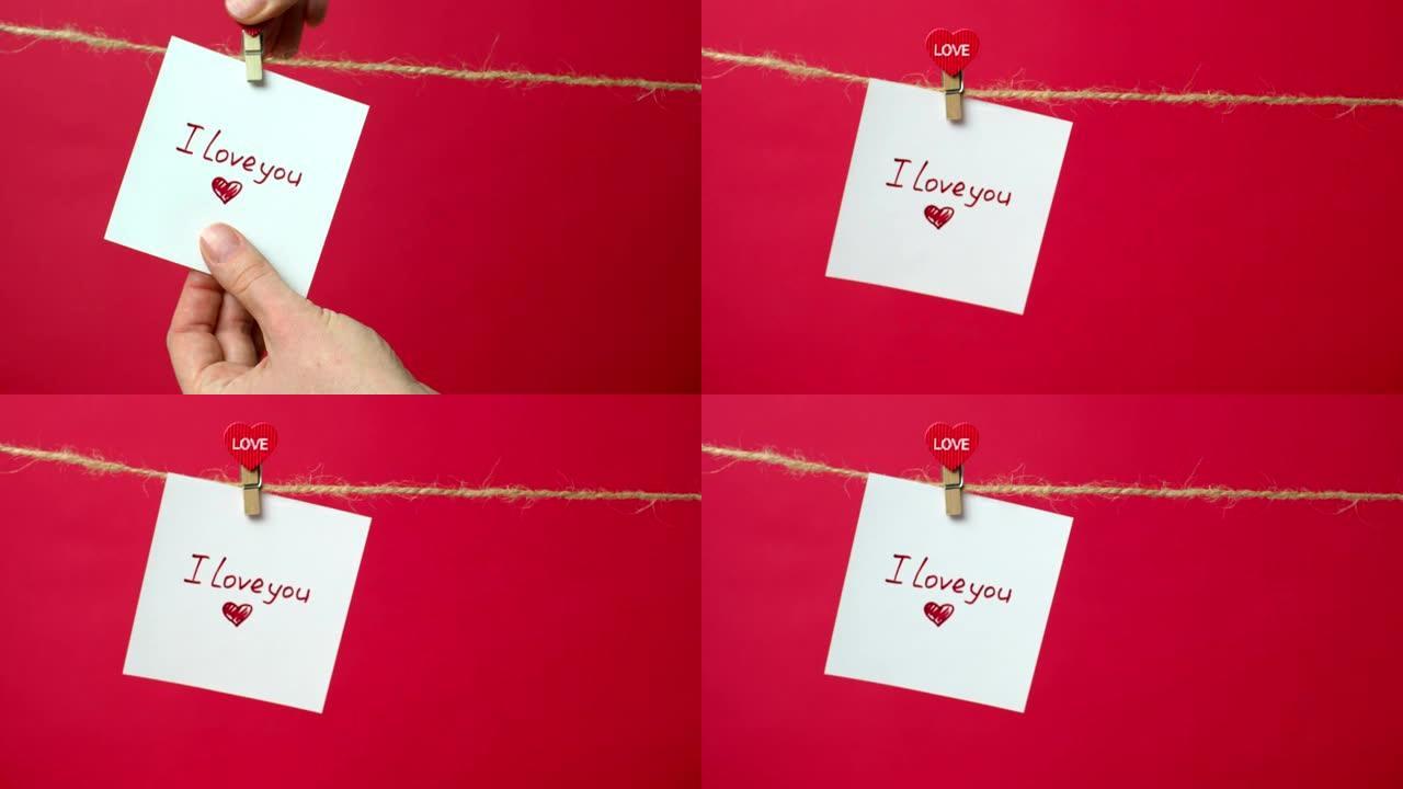 绳子上写着 “我爱你” 文字的特写。女孩的手将一张白纸挂在衣夹上的红色背景上。爱情宣言、婚礼、情人节