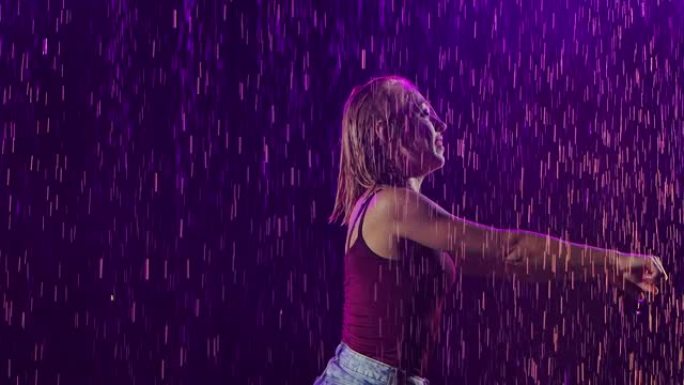 一个穿着短裤和t恤的苗条金发女郎在倾盆大雨中享受着萨尔萨舞。年轻热情的女人在工作室里以紫光和烟雾为背