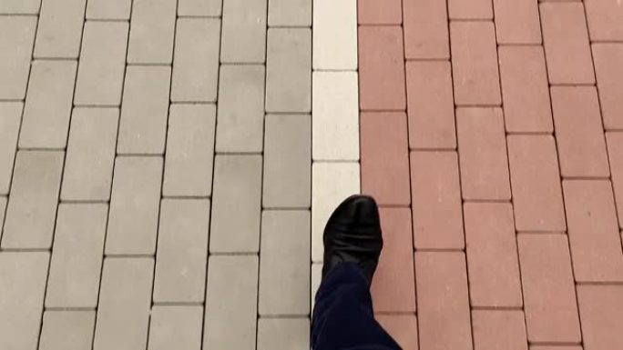 一个男人沿着石路行走的腿的顶部的特写镜头。一名穿着黑色鞋子的男子沿着人行道的白线行走，在灰色和红色的