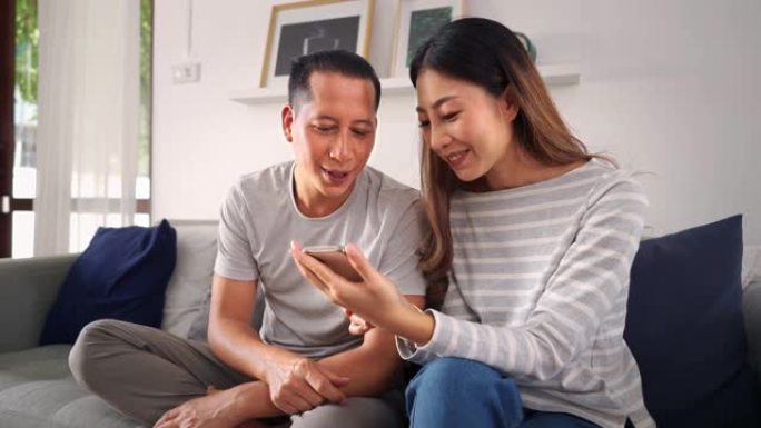 30多岁的亚洲年轻人一起玩手机的男人和女人坐在家里舒适的客厅的沙发上。幸福的夫妇互联网用户穿着休闲服