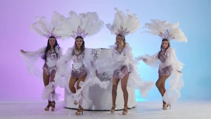 一群可爱的女人穿着白雪皇后的服装和羽毛头饰在一个大蛋糕旁跳舞。舞者在浅蓝色的工作室背景下摆姿势。新年
