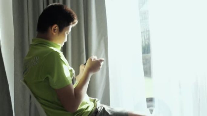 亚洲男孩坐在家里窗户附近的椅子上玩智能手机在线游戏