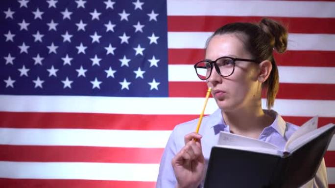 戴眼镜的学生女孩困惑地思考，并在美国国旗背景的笔记本上写下想法。美国学生，美国学生。美国的教育
