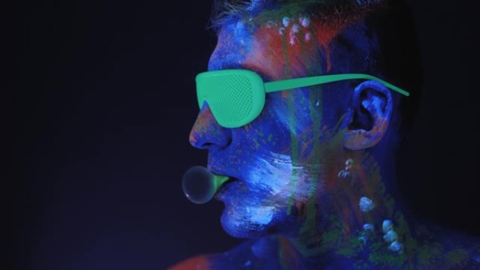 一个戴着绿色氖气眼镜画着紫外线画的男人正在吹泡泡糖。