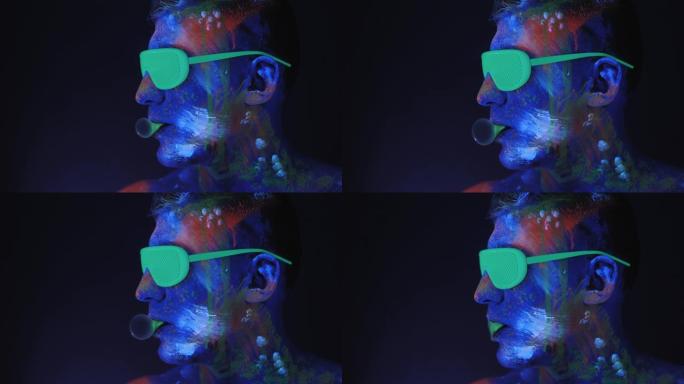 一个戴着绿色氖气眼镜画着紫外线画的男人正在吹泡泡糖。