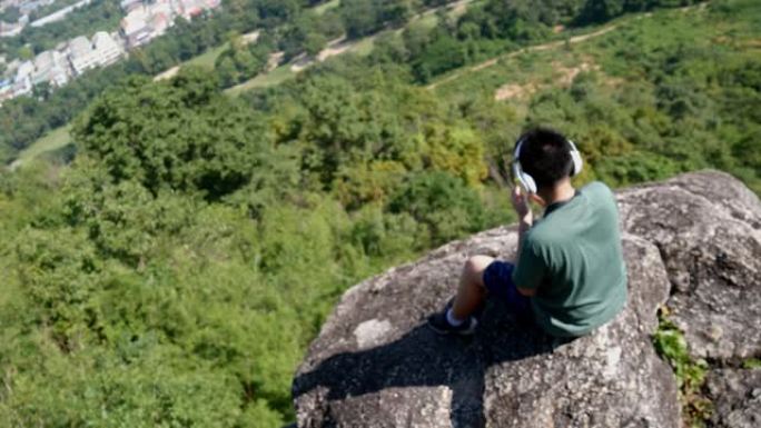 在阳光明媚的日子里，带着自然背景，模糊的亚洲男孩在山顶上戴着耳机听音乐。