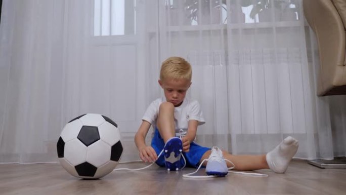 一个小男孩坐在地板上，学习将鞋带系在足球靴上。