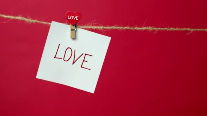 用文字爱在绳子上的音符特写。女孩的手将一张白纸挂在衣夹上的红色背景上。爱情宣言、婚礼、情人节的概念。