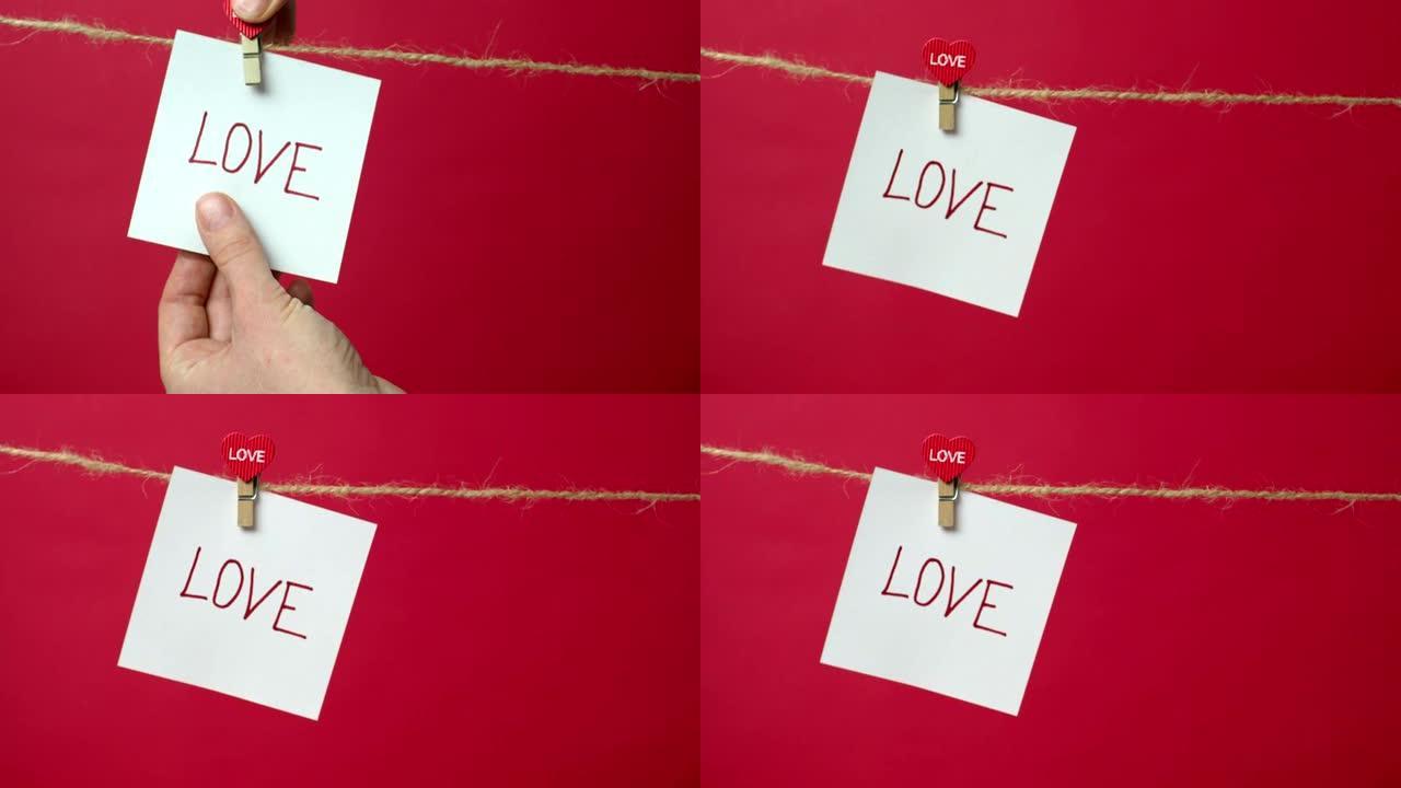 用文字爱在绳子上的音符特写。女孩的手将一张白纸挂在衣夹上的红色背景上。爱情宣言、婚礼、情人节的概念。