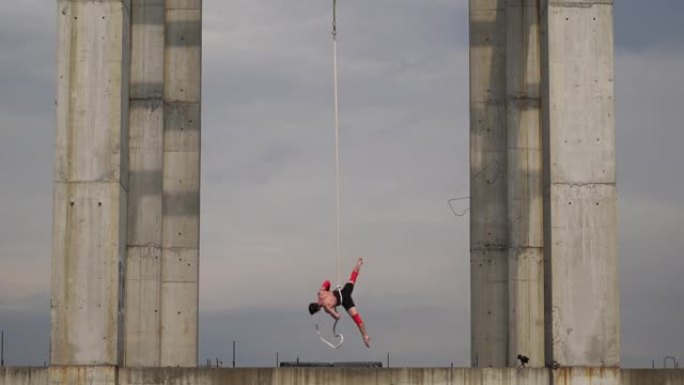 强壮肌肉的马戏团艺术家在空中绳索上表演天空背景上的两个混凝土柱之间的室外，力量、风险和危险的概念