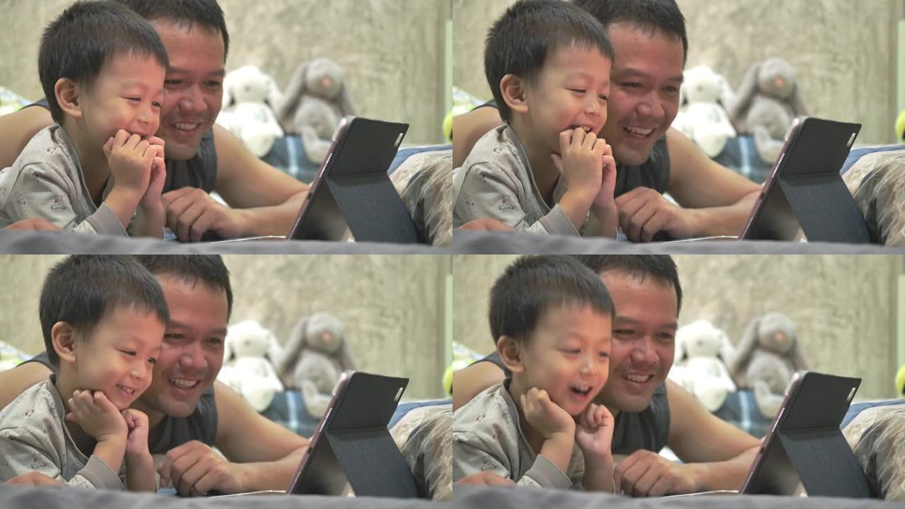 亚洲父子正从卧室的平板电脑上愉快地观看动画片。