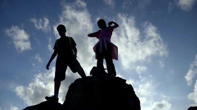 剪影男孩和女孩站在山顶，举手与蓝天，成功的概念。
