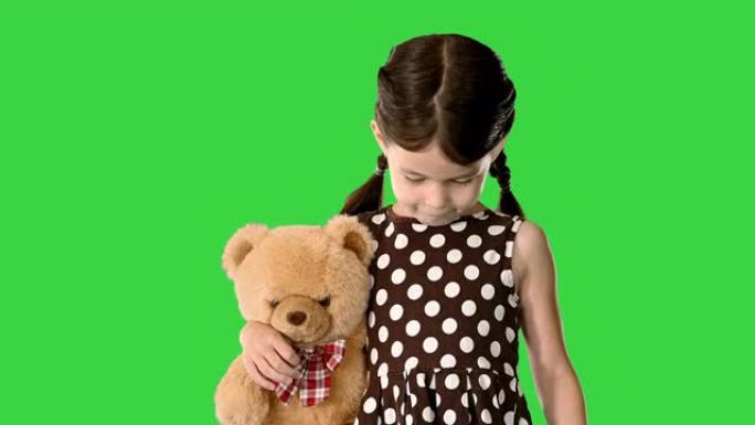 穿着圆点连衣裙的害羞小女孩在绿色屏幕上走路时抱着泰迪熊，低头看着她的脚，色键