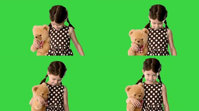 穿着圆点连衣裙的害羞小女孩在绿色屏幕上走路时抱着泰迪熊，低头看着她的脚，色键