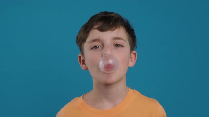 快乐的男孩嚼口香糖，学习膨胀气泡并使气泡破裂，快乐地微笑着看着蓝色背景上的相机慢动作。孩子。童年。人