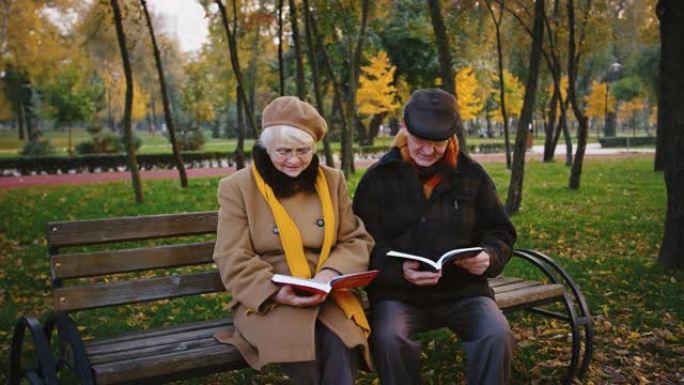 戴着眼镜和保暖外套的退休夫妇坐在秋天公园的木凳上看书