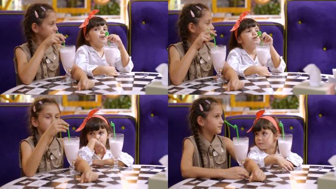 儿童喝牛奶鸡尾酒。两个四岁和七岁可爱的微笑女孩坐在比萨店的桌子旁，喝着眼镜上的奶昔。慢动作50 FP