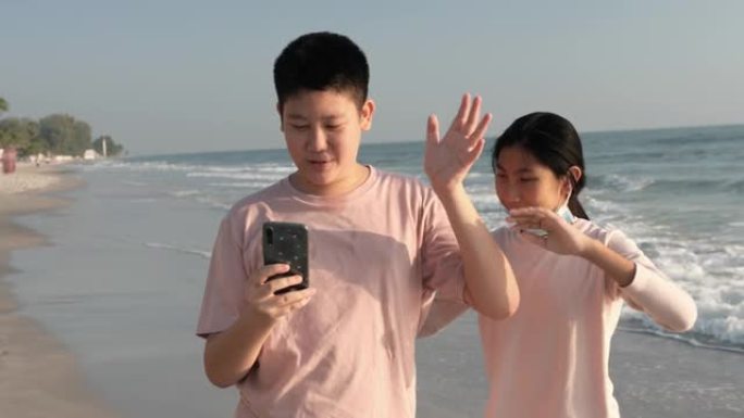 快乐的亚洲男孩和他的妹妹在假期，科技概念早上在海滩上通过智能手机与家人拍照自拍或视频通话。
