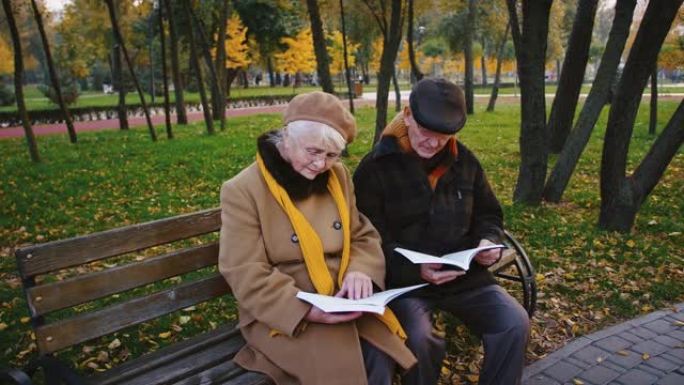 一副戴眼镜和保暖外套的退休眼镜坐在秋天公园的木凳上看书和说话