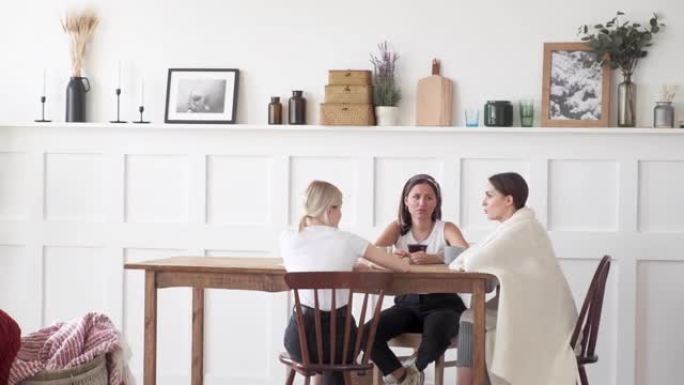 三个女孩坐在家里一个大房间的桌子和椅子上聊天，互相讲故事