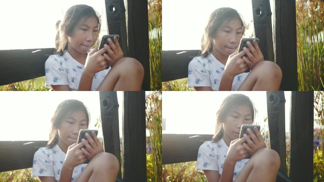 快乐的亚洲女孩坐在木椅上，使用智能手机与太阳灯背景，技术概念。