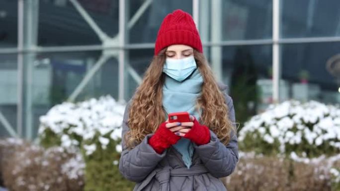 戴着防护医用口罩的年轻卷发妇女走上街头使用电话短信滚动浏览互联网搜索新闻covid19冠状病毒病毒保