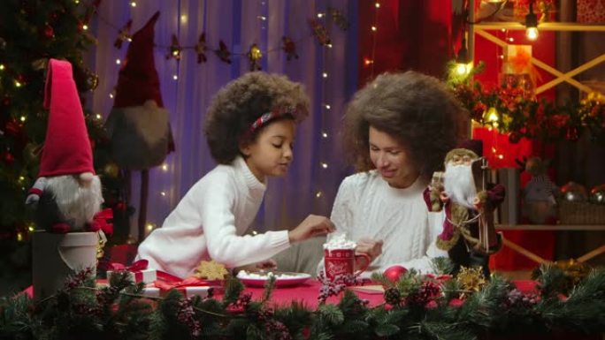 非裔美国妈妈和小女儿用鲜奶油和棉花糖装饰一杯热饮料。快乐全家福，新年庆祝概念