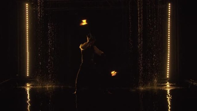 雄性驯火师站在雨滴之间时，在铁链上旋转燃烧的火球。晚上在黑暗的工作室里精彩的火灾表演。烟雾中的轮廓。