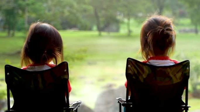 两个女孩坐在躺椅上享受乡村露营假期的早餐。家庭在大自然和户外活动中放松时间的概念。