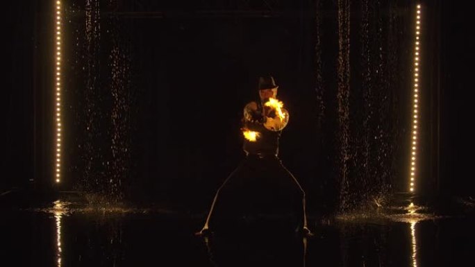 雄性驯火师站在雨滴之间时，在铁链上旋转燃烧的火球。晚上在黑暗的工作室里精彩的火灾表演。烟雾中的轮廓。