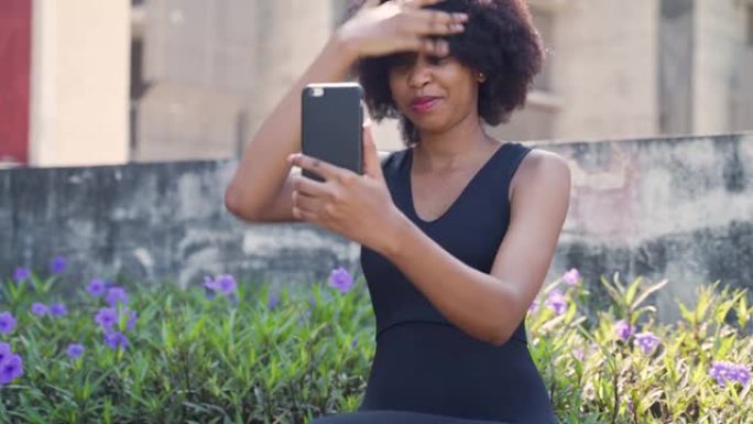 年轻的非洲裔美国妇女用手机看着自己，修理她的非洲头发。女性黑色慢跑者坐在室外公园里，使用手机摄像头。