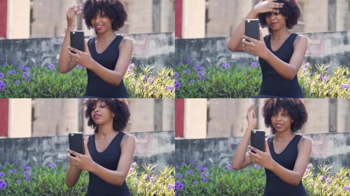 年轻的非洲裔美国妇女用手机看着自己，修理她的非洲头发。女性黑色慢跑者坐在室外公园里，使用手机摄像头。