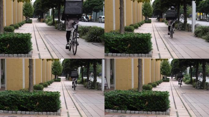 带着黑色背包和防护面具的年轻人快递员在街上骑着自行车穿过城市，运送食物。戴防护头盔的人。视频是从后面
