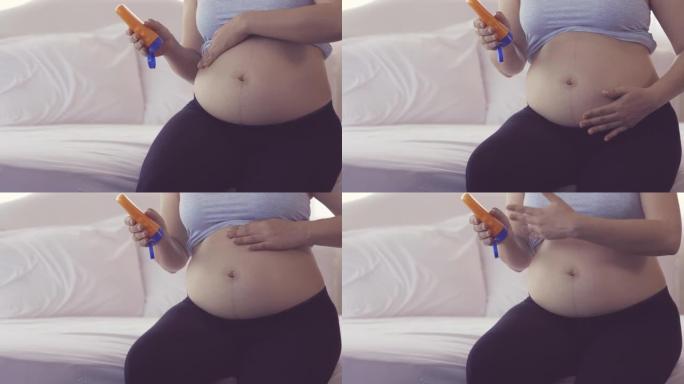 年轻的亚洲孕妇母亲坐在床上时拿着乳液瓶和腹部擦乳液