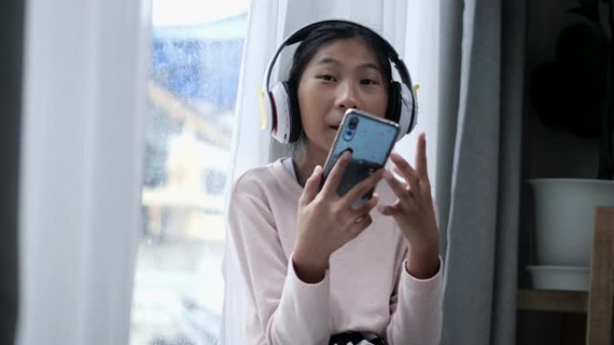亚洲人坐在家里窗户附近的地板上，使用智能手机和耳机看电影。