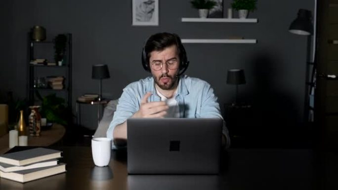 一位年轻的男性高管在家里舒适的房间里发誓，使用笔记本电脑，通过耳机与朋友或客户在线交谈