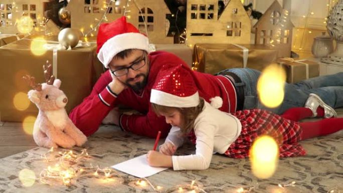 幸福的家庭，年轻的父亲和戴着红色圣诞老人帽子的小女孩在圣诞树的背景上给圣诞老人写信。圣诞节和新年
