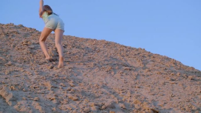 快乐的女孩少年在蓝天背景上攀登山顶沙山。低角度视角快乐少女在夏季沙漠的高沙堆上。